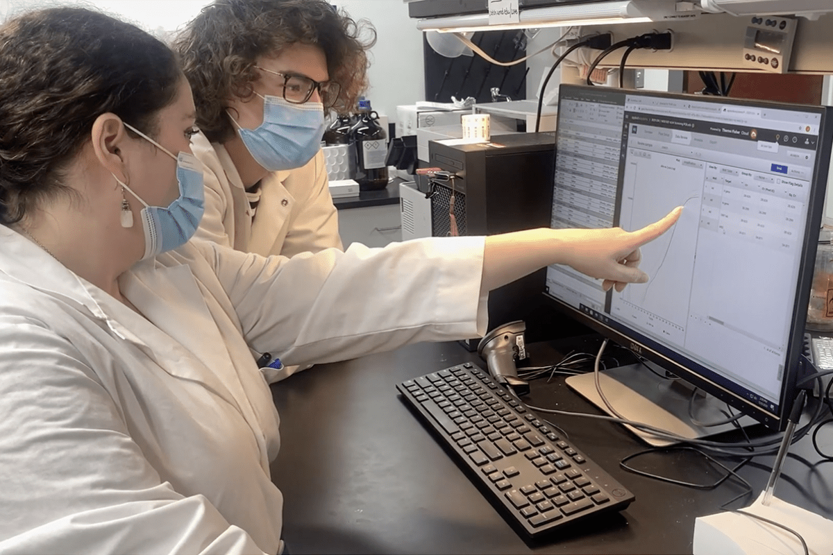 Two people wearing masks and white lab coats look at data on computer screen, one points to detail on screen