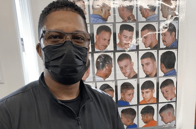 Barber wearing a mask in his shop with Haircut diagram behind him